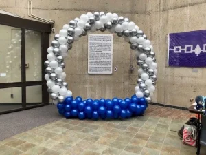 A large balloon arch with blue, silver, and white balloons stands in a building lobby near posters and glass doors.