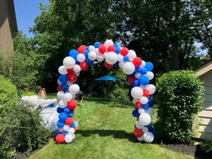 A red, white, and blue balloon arch stands on a grassy lawn with trees and bushes in the background.