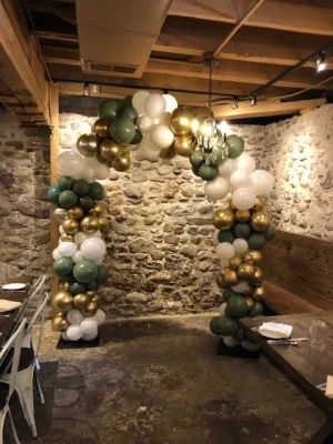 Balloon arch in green, white, and gold stands in a rustic room with stone walls and wooden ceiling beams.