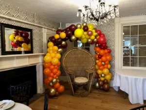 A wicker chair sits under a colorful balloon arch in a decorated room with a chandelier and patterned walls.