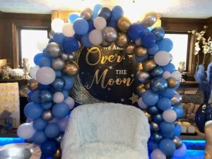 Blue and silver balloon arch frames a sign reading "Over the Moon" behind a white chair in a decorated room.