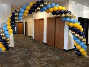 Balloon arch with blue, yellow, and gold balloons stands in front of wooden doors in an indoor hallway.