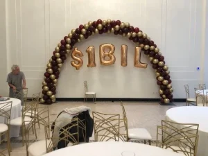 Balloon arch with gold "$1BIL" balloons in the center; empty chairs and tables arranged around the room.