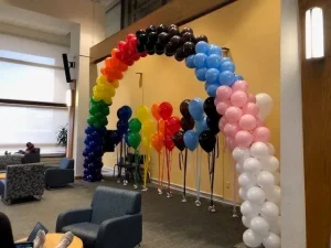 Balloon arch in various colors stands in a lounge area with chairs, tables, and a person sitting in the background.