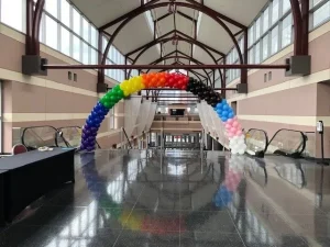 A rainbow balloon arch stands in a large, empty hallway with escalators and tall windows.
