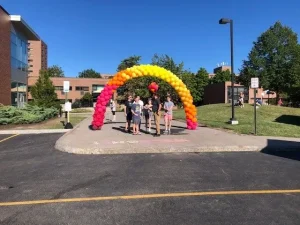 Six people stand under a rainbow balloon arch on a sunny day outside a building, with greenery in the background.