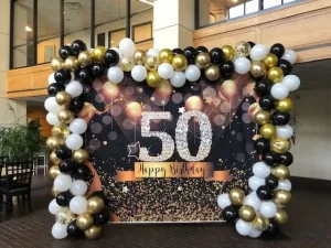 Balloon arch with a "50 Happy Birthday" banner in gold, black, and white in a decorated indoor space.