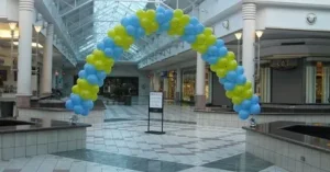 A blue and yellow balloon arch stands in an empty shopping mall corridor near a sign.