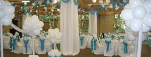 A banquet hall decorated with white and blue balloons, round tables, and chairs covered in white with blue bows.