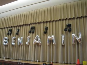 Silver balloons spelling "BENJAMIN" and black musical note balloons are attached to beige stage curtains.