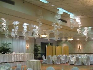 Banquet hall with white balloons hanging from the ceiling, round tables, and chairs covered in white fabric.