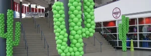 Green balloons arranged in the shape of cacti stand near stairs inside a building with a sports shop.