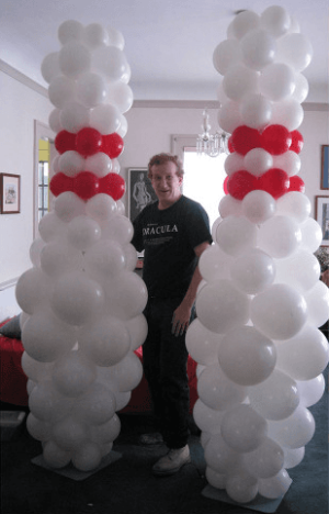 A person stands between two tall balloon sculptures resembling bowling pins, made of white and red balloons.
