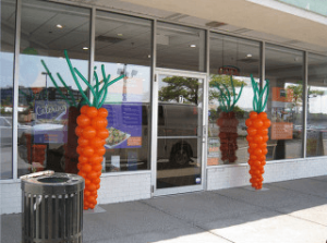 Large carrot-shaped balloon decorations are displayed outside the entrance of a storefront with glass windows.