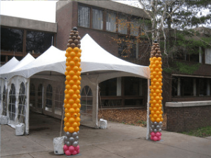 Two tall balloon columns resembling pencils stand outside a white event tent near a brick building.