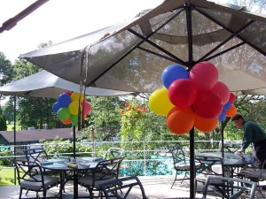 Outdoor patio with tables, chairs, umbrellas, and colorful balloon clusters; person stands near one table.