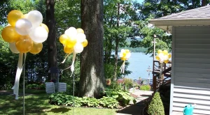 Yellow and white balloon clusters decorate a lakeside yard with trees, shrubs, and a partial building in view.