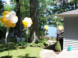 Yellow and white balloon clusters on poles decorate a shaded backyard overlooking a lake on a sunny day.