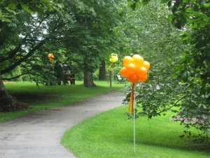 Orange and yellow balloons are tied to sticks along a paved park path surrounded by green grass and trees.