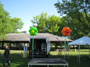 Outdoor stage with a microphone, green and orange balloon decorations, a truck, and a picnic shelter in the background.