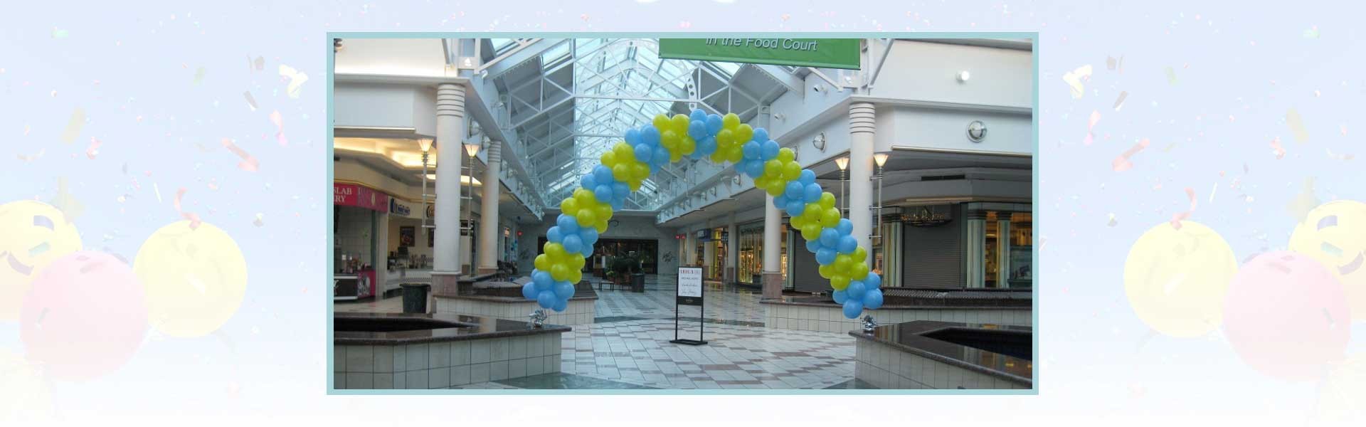 A blue and yellow balloon arch stands in the center of an indoor shopping mall with shops in the background.