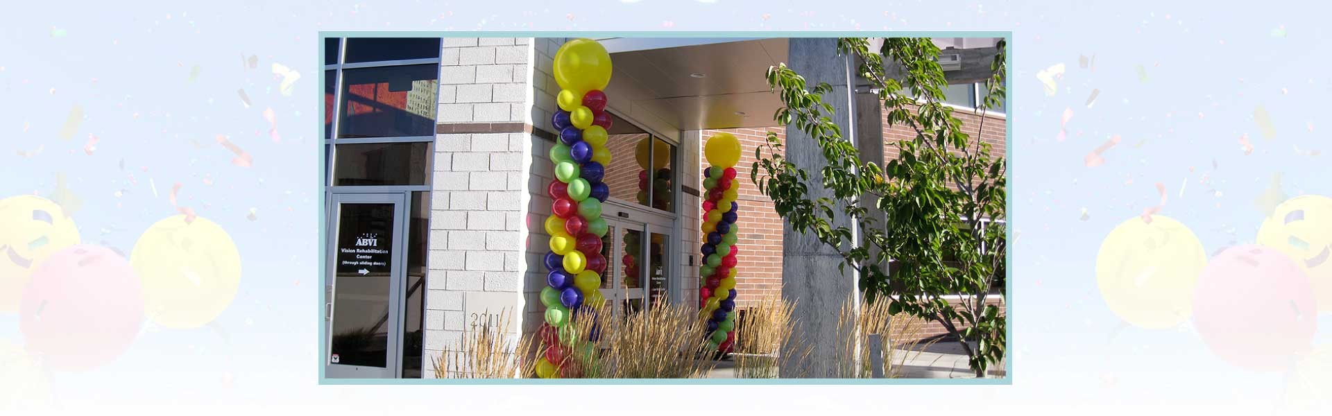 Two columns of colorful balloons decorate the entrance of a modern brick and glass building on a sunny day.
